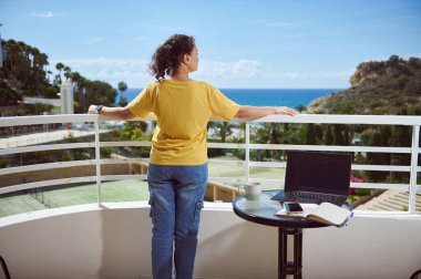 Diz üstü bilgisayarı ve kahvesiyle balkondan okyanus manzarası seyreden bir kadın. Rahatlama, uzak çalışma ve huzurlu kıyı yaşam tarzı kavramı.
