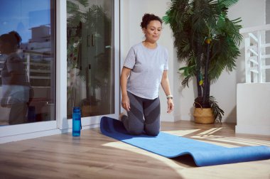 Bir kadın kapalı bir alanda yoga minderinde egzersiz yapmaya hazırlanıyor. Etrafında bitkiler ve su şişesi var. Sağlık, sağlık ve farkındalığı ön plana çıkarıyor..
