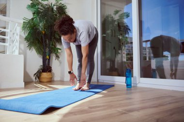 Genç bir kadın balkondaki mavi yoga minderinde geriniyor. Yakınlarda bir su şişesi var. Cam kapılardan yansıyan yansıma, bitki dekoru ile sakin atmosferi güçlendiriyor..