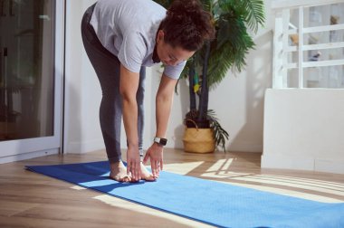 Odaklanmış bir kadın yoga egzersizleri yapar, yoga minderinde durur ve öne doğru eğilir. Kapalı alan parlak ve saksı bitkileriyle süslenmiş, dingin bir atmosfer yaratıyor..