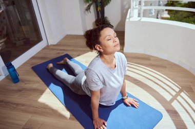 Huzurlu bir kadın güneş ışığıyla aydınlatılmış bir balkonda yoga yapıyor. Atmosfer sakin, rahatlama ve refahı teşvik ediyor..