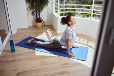 Bir kadın, güneş ışığıyla aydınlatılmış bir alanda mavi bir egzersiz minderinin üzerinde gerinerek yoga yapıyor. Huzurlu sahnede bir su şişesi ve refah aktiviteleri için ideal doğal ışık bulunuyor..