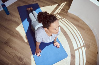 Yoga yapan bir kadın, güneş ışığıyla aydınlatılmış bir alanda, ahşap döşemeli ve sakin atmosferli bir yerde, sağlığını ve farkındalığını vurgulayarak yoga pozu veriyor..