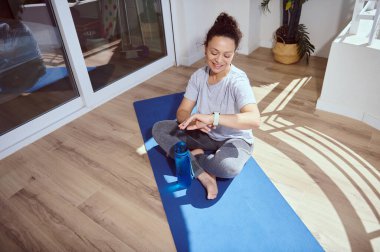 Bir kadın, güneşli bir odada yoga minderinde oturur, egzersizden sonra akıllı hareketlerini kontrol eder, rahatlama, zindelik ve farkındalık gösterir..