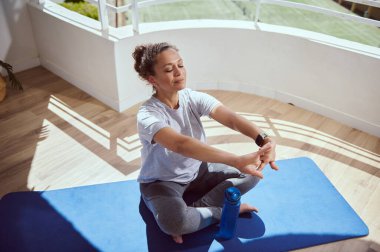 Yoga minderinde bacak bacak üstüne atmış bir kadın, iyi aydınlatılmış bir balkon ortamında meditasyondan zevk alıyor, sakinlik ve farkındalık saçıyor..