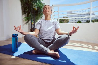 Açık havada meditasyon yapan bir kadın mavi bir minderde huzur içinde yoga yapıyor. Huzur ortamında sağlık, farkındalık ve zihinsel rahatlık vurgulanıyor..