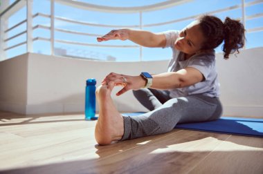 Bir kadın, kapalı bir alanda yoga minderinde fitness esneme hareketi yapıyor. Parlak çevre, fiziksel sağlık ve refahı korumak için odaklanmayı ve kendini adamayı teşvik eder..