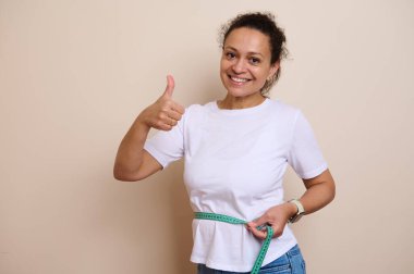 Günlük beyaz gömlekli gülümseyen kadın, kişisel fitness hedeflerine ulaşmadaki ilerlemesinden duyduğu heyecanı ve memnuniyeti ifade eden bir bant ile belini ölçüyor. Sağlıklı bir yaşam tarzı.