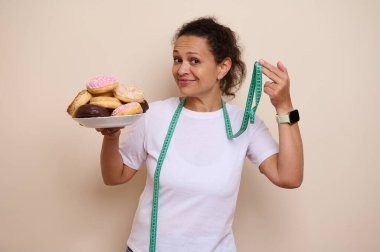 Neşeli bir kadın elinde bir tabak renkli çörek tutarken ölçüm bandıyla el kol hareketi yaparak hoşgörü ve sağlıkla ilgili kararlar arasındaki dengeyi sembolize ediyor..