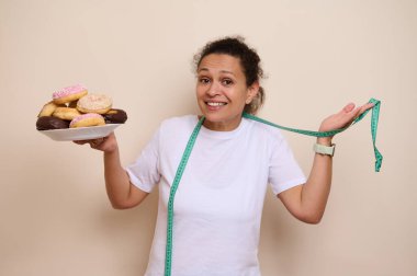 Neşeli bir kadın, tatlı hoşgörü ve sağlık arasında denge kurar. Elinde lezzetli bir tabak çörekle ilerlemesini mezura ile ölçer. Denge kavramını betimler..