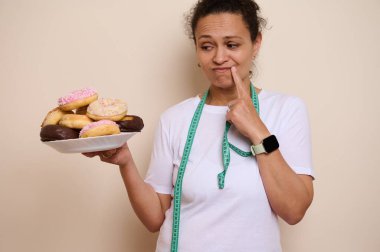 Düşünceli bir kadın, sağlığını kucaklarken şekerli çöreklere dalmayı düşünür, mezura ve akıllı saatiyle sembolize edilir, hoşgörü ve sağlığı dengeleyen bir yaşam tarzını gösterir..
