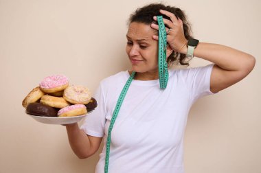 Bir kadın sağlık kararlarını düşünürken bir tabak donut tutuyor, hoşgörü ve sağlıklı yaşam tarzına zıt düşüyor, seçenekleri, diyet sorunlarını ve Ulusal Sağlık Ayı temaları için mükemmel..