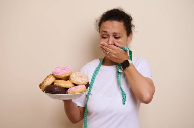 Bir kadın elinde rengarenk donut tabağıyla dikiliyor, düşünceli görünüyor. Hoşgörü ve sağIık arasında yol alırken boynunda bir mezura asılı duruyor..