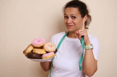 Bir tabak renkli çöreği düşünen bir kadın, hoşgörü ve sağlık arasındaki dengeyi vurguluyor, Ulusal Sağlık Ayı ve Kremalı Çörek Günü temaları.