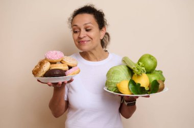 Elinde meyve ve donut tabakları tutan bir kadın sağlıklı beslenmeye hoşgörü ile zıt düşüyor, refahı ve yaşam dengesini yansıtıyor..
