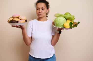 Beyaz bluzlu bir kadın iki tabak alır, biri canlı renkli tatlı çöreklerle, diğeri taze meyve ve sebzelerle dolu, tatlı ve sağlıklı yemek arasında bir seçim yapmayı düşünür..