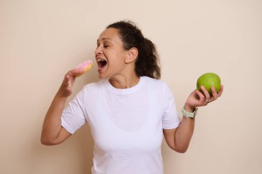 Pembe jöleli donut ve yeşil elma tutan bir kadının canlı tasviri hoşgörü ile sağlıklı beslenme arasındaki dengeli kararı gösteriyor..