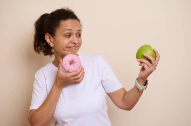 Pembe buzlu donut ile taze yeşil elma arasında seçim yapan bir kadın hoşgörü ve sağlıklı beslenme alışkanlıklarını sembolize eder..