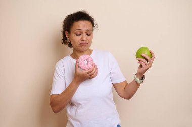 Bir kadın yeşil elma ve pembe çörek arasında tartışıyor. Görüntü, çağdaş bir ortamda sağlık, cazibe ve yaşam tarzı kararlarını iletir..
