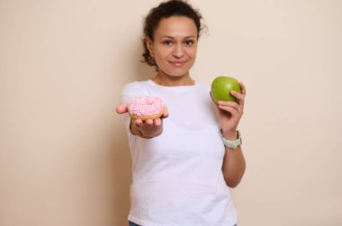 Bir kadın hem sağlıklı bir elma hem de cazip pembe bir çörek göstererek dengeli bir yaşam tarzı sergiler. Seçenekleri, tercihleri ve refah ve hoşgörü unsurlarını vurgulamak.