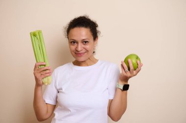 Neşeli bir kadın taze yeşil bir elma ve bir demet kereviz sapı tutarak dengeli gıda seçeneklerini ve sağlığını temsil ederek sağlıklı bir yaşamı teşvik eder..