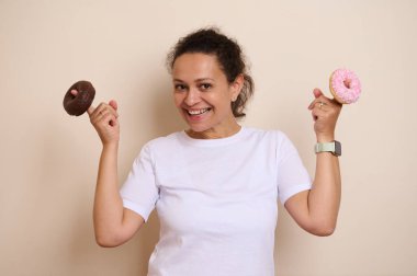 Neşeli bir kadın elinde çikolata ve pembe donut tutarken gülümsüyor. Tatlı ikramlar ve mutluluklar sunan bu görüntü, hoşgörü, haz ve özel anları tatlılarla kutluyor..