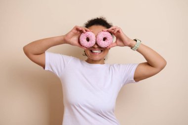 Neşeli bir kadın iki pembe çöreği gözünün önünde tutuyor, neşeli ve neşeli bir şekilde poz veriyor. Görüntü pozitif ve yaratıcı bir zevk yayar..