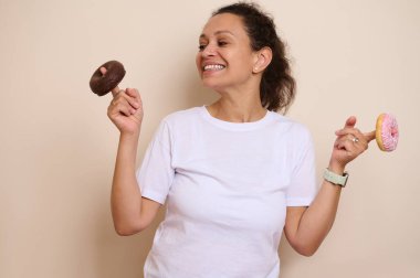 Beyaz tişörtlü neşeli yetişkin bir kadın iki renkli çörek tutmaktan ve gülümsemekten zevk alır. Tarafsız arka plan canlı donut renklerini güçlendiriyor, dinamik ve davetkar bir görsel yaratıyor.