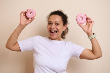 Neşeli bir kadın beyaz bir gömlek giyer, elinde iki pembe donut tutarken parlak bir şekilde gülümser, günlük bir ortamda neşe ve canlılık yayar. Yemek, eğlence ve yaşam tarzı temaları için mükemmel.