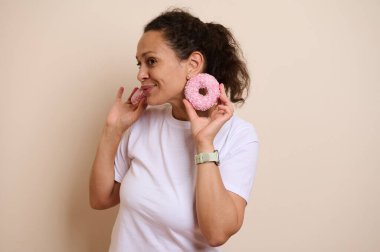 Neşeli bir kadın elinde parlak pembe donutlar tutarken gülümsüyor, neşeli ve mutlu bir sahne yaratıyor. Görseller neşe, renk ve hoşgörü vurguluyor.