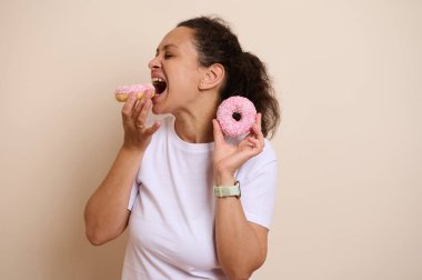 Neşeli kadın pembe krema ve şekerle kaplı taze pişmiş donutların tadını çıkarıyor. Keyifli ve neşeli bir ifadeyle hoşgörü ve tatlılığı vurgulayan lezzetli bir an..