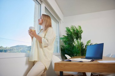 Rahat bir kadın ev ofisinin penceresinin yanında durup elinde bir fincan tutarak dışarı bakıyor. Havadar, minimalist çalışma alanı bir dizüstü bilgisayar, defter ve yeşillik içeriyor, sükunet ve verimliliği yansıtıyor..