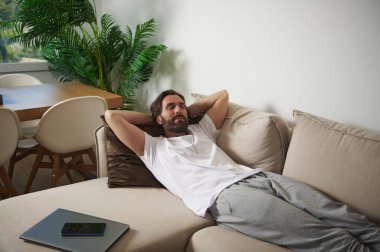 Modern mobilyalar ve yeşil bitkilerle çevrili nötr bir koltukta uzanan bir adamla huzurlu bir yuva kurmak, rahatlama ve rahatlığın özünü somutlaştırıyor..