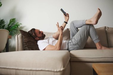 Günümüz minimalist oturma odasında bej bir koltukta rahat bir şekilde uzanan, akıllı telefonuyla meşgul olan, rahatlama ve dijital etkileşimi ileten, günlük kıyafetli bir adam..