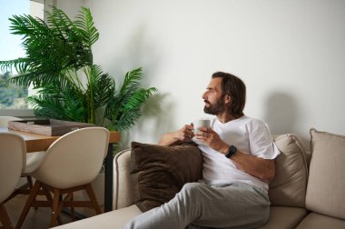 Görüntü, rahat bir oturma odasında oturmuş, bir fincan kahveyle huzurlu bir anın tadını çıkaran, kapalı alan bitkileri ve minimalist dekor ile tamamlanmış bir bireyi sergiliyor, rahatlamayı öneriyor..