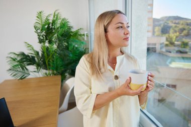 Aydınlık bir ofisin penceresinin yanında duran dingin bir kadın ev bitkileri ve sıcak bir atmosferle rahatlama ve ilham verici anlar yaşıyor. Ev rahatlığı ve sükunet kavramları için ideal..