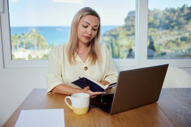 Odaklanmış bir kadın şık bir ev ofisinden, manzaralı bir kıyı manzarasına bakan, dijital göçebeliği ve iş hayatı dengesini gösteren uzak bir işe girişiyor..