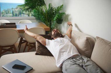 Adam, modern, sakin bir oturma odasında rahat bir dekor ve canlı bitkilerle kanepeye uzanıyor. Rahatlık ve huzur dolu bir anın tadını çıkarıyor..