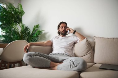 Bej bir kanepede rahat bir şekilde oturan ve telefon konuşmasına odaklanan bir adam. İçi kapalı bitkiler ve minimalist mobilyalar içeriyor. Huzurlu bir ev ortamı yaratıyorlar..
