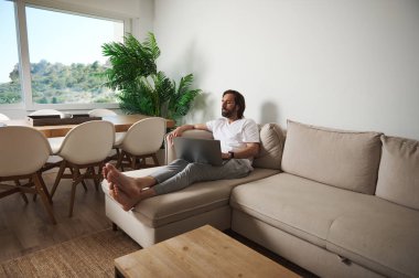 Çağdaş bir ev ortamında dizüstü bilgisayarla çalışırken rahat bir kanepenin üzerinde minimalist bir dekor ve doğal ışık barındıran bir anın tadını çıkarıyor..