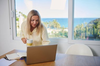 Kendini işine adamış bir kadın, elinde bir kahve fincanı tutarken güneşli bir günde okyanus manzarasına bakan minimalist bir ev ofisinde çalışıyor..
