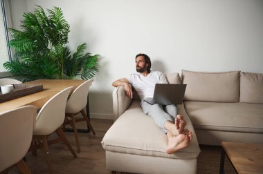 Bir adam modern bir kanepede minimalist, rahat bir iç mekanda rahat bir şekilde uzanırken rahat üretkenliği sembolize eden bitkiler ve doğal ışığı temsil eder..