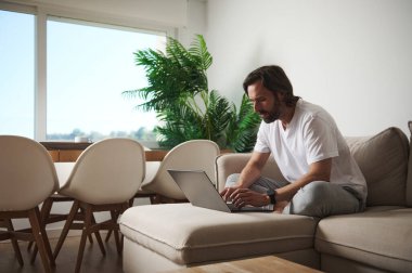 Odaklanmış bir adam, üretkenlik ve rahatlığı vurgulayan, minimalist dekorlu, parlak, modern bir oturma odasında dizüstü bilgisayar kullanır. Rahat bir çalışma alanında ideal boş zaman.