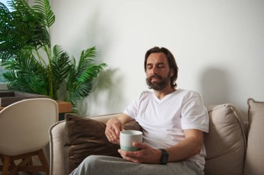 Minimalist bir oturma odasındaki koltukta rahat bir şekilde oturan, elinde bir kupa tutan ve ev bitkileri ve rahat mobilyalarla dolu huzurlu bir anın tadını çıkaran bir birey..