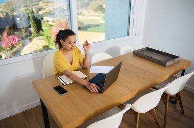 Tahta bir masanın üzerinde dizüstü bilgisayarı olan, geniş pencerelerden görülebilen sakin bir çevreyle çevrili minimalist bir ev ofisinden çalışan kişi, uzaktan çalışma ve dijital bağlantıyı vurguladı.