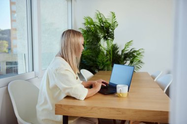 Ev ofisinin ahşap masasında oturmuş dizüstü bilgisayar kullanan bir kadın, minimalist bir çalışma alanı ve doğal ışık gösteriyor..