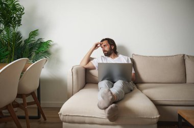 Uzaktan çalışan profesyonel bir adam rahat bir oturma odasında dizüstü bilgisayarla fikirleri yansıtıyor. Etrafı yeşillik ve mobilyalarla çevrili, sakin bir telekomünikasyon ortamı yaratıyor..