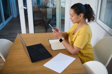 Konforlu bir minimalist ev ofisinde uzaktan çalışan profesyonel kadın, teknolojiyi ve görevleri verimli bir şekilde dengelemekte, modern serbest ve girişimci yaşam tarzını somutlaştırmaktadır..