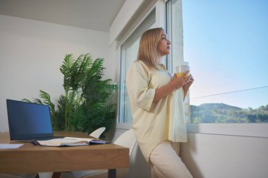 Bir kadın şık bir ev ofisinin penceresinde elinde fincanla dikiliyor ve açık hava manzarasının tadını çıkarıyor. Çalışma alanı dekor rahatlama ve üretkenlik iletir.
