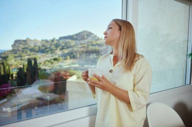 Sakin bir atmosferde, bir kadın evin içinde içecekten zevk alır, büyük cam bir pencereden manzarayı seyreder. Rahatlamayı, sakinliği ve huzurlu yaşamı temsil eder..
